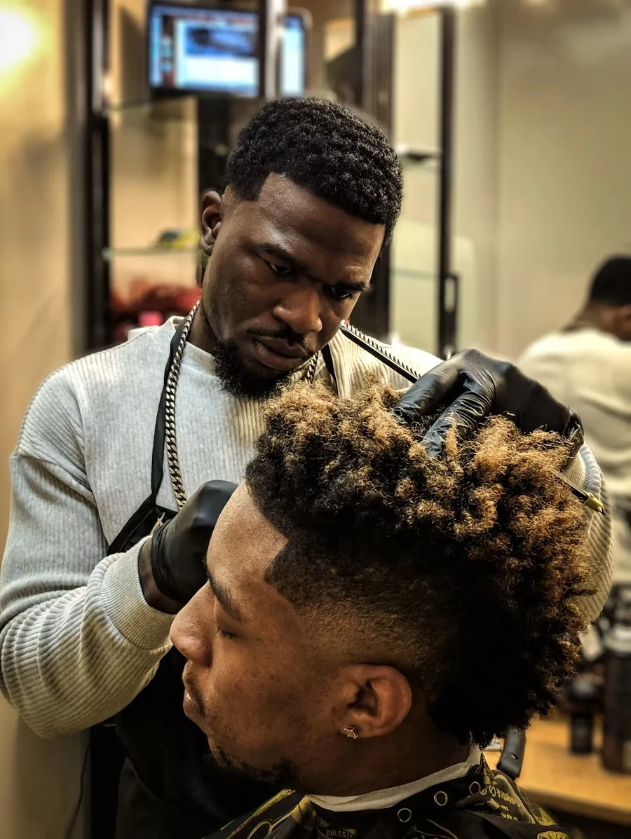 A barber in black gloves shaping a client's curly hair with clippers, with a fade along the side of the head