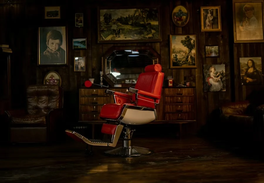 A red leather barber chair in a dark wood-panelled room hung with framed paintings, leather armchairs behind
