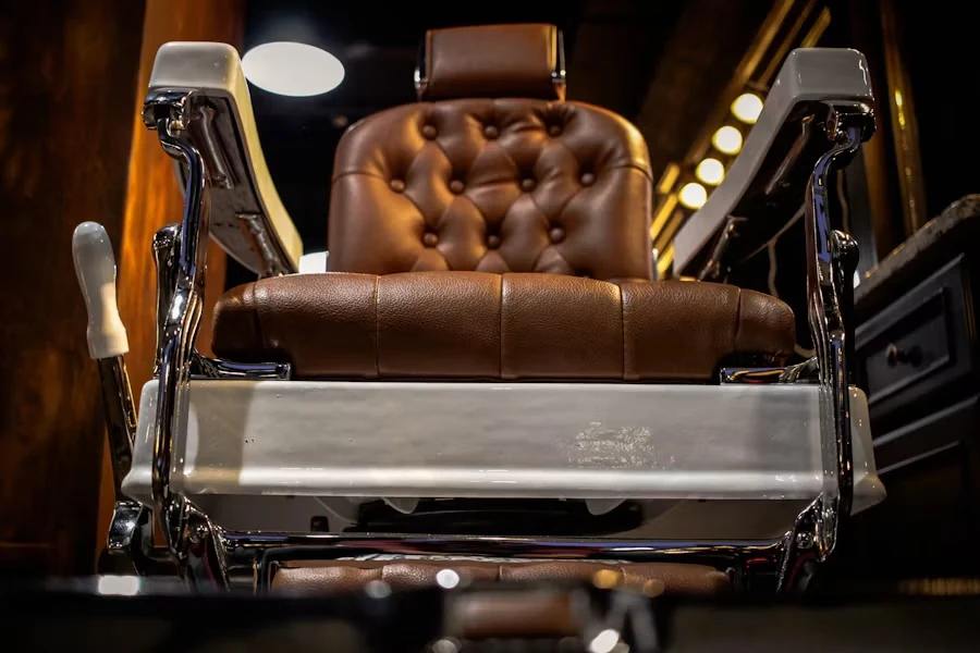 Low-angle view of a vintage barber chair with tufted brown leather, chrome frame and white armrests in a dimly lit shop