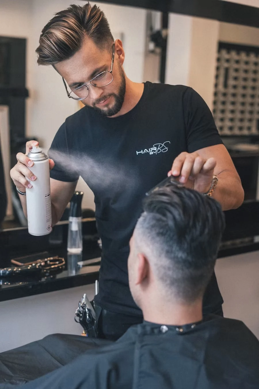 A barber in glasses and a black t-shirt spraying hairspray while styling a seated client's hair