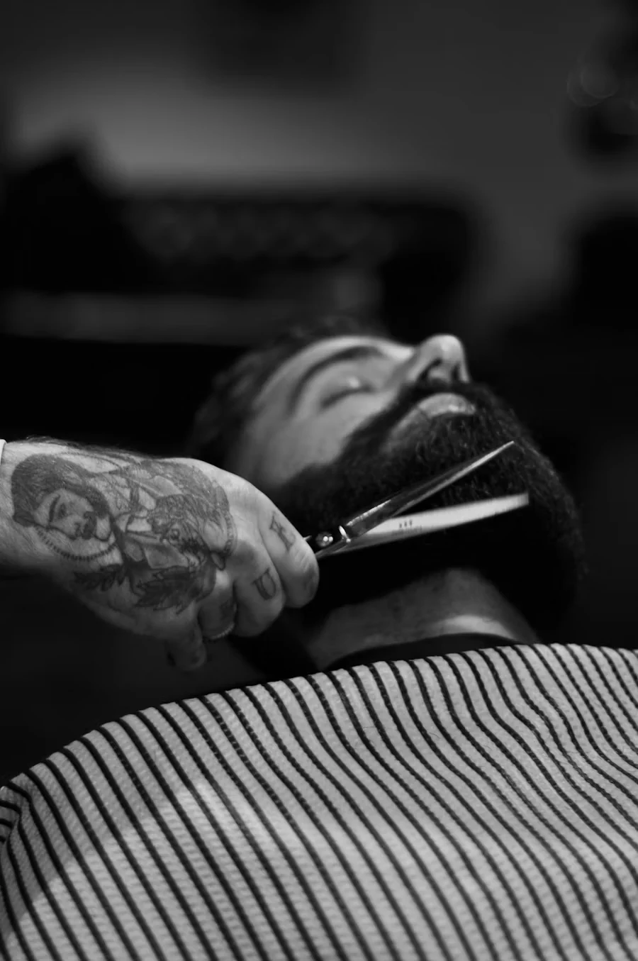 Black and white photo of a tattooed barber trimming a client's beard with scissors, the client lying back under a striped cape