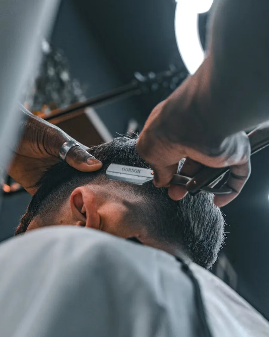 Close-up of a barber's straight razor shaping the neckline of a skin fade on a client wearing a grey cape