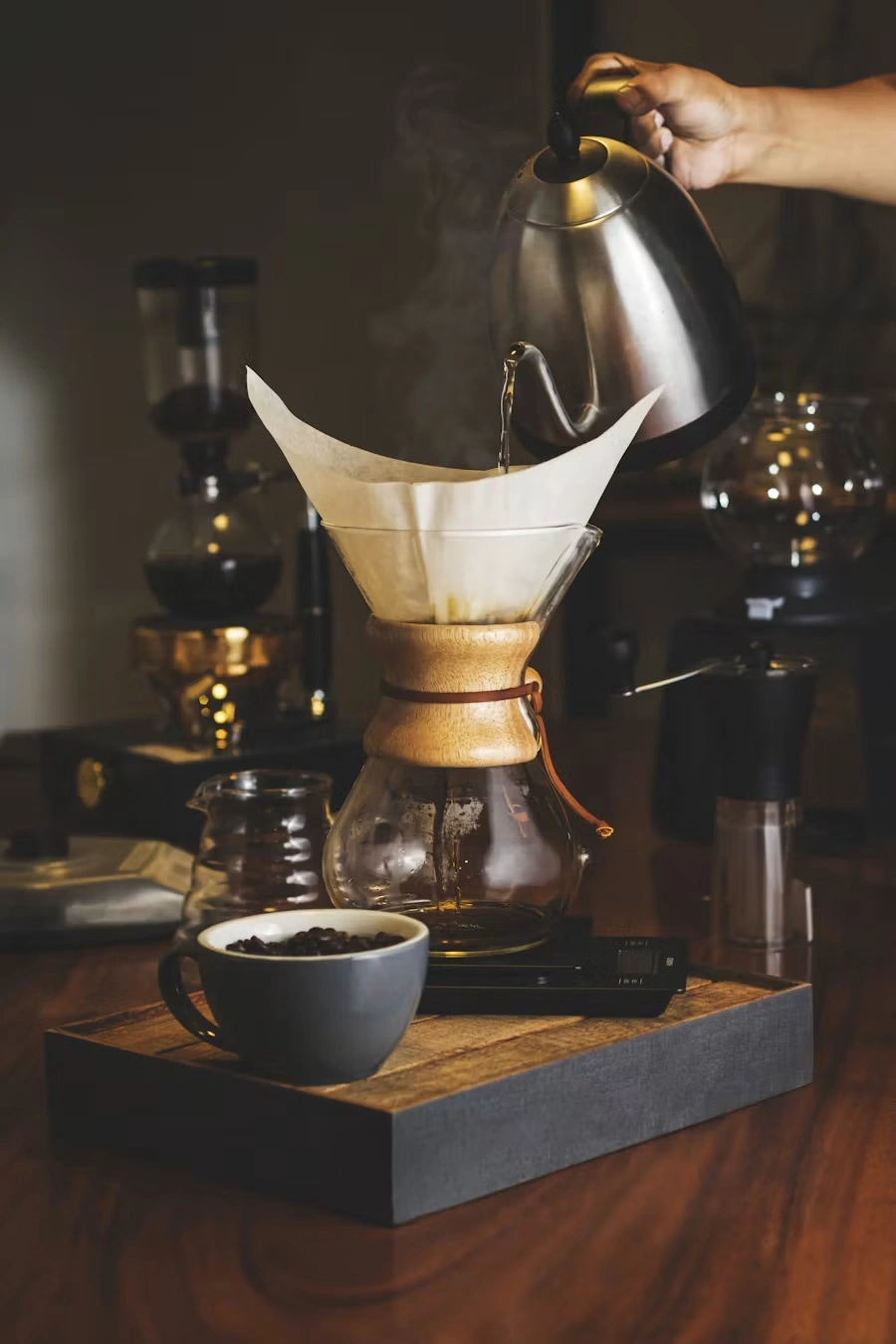 A hand pouring hot water from a gooseneck kettle over a paper filter in a Chemex, with a cup of coffee beans beside it