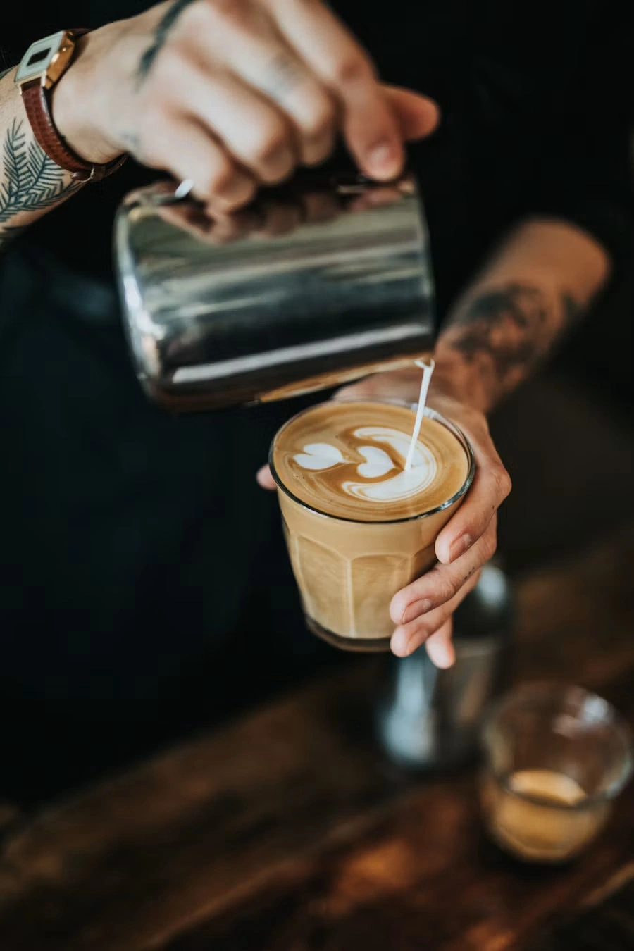 A barista pouring steamed milk from a steel jug into a glass of coffee, making a heart-shaped latte art pattern
