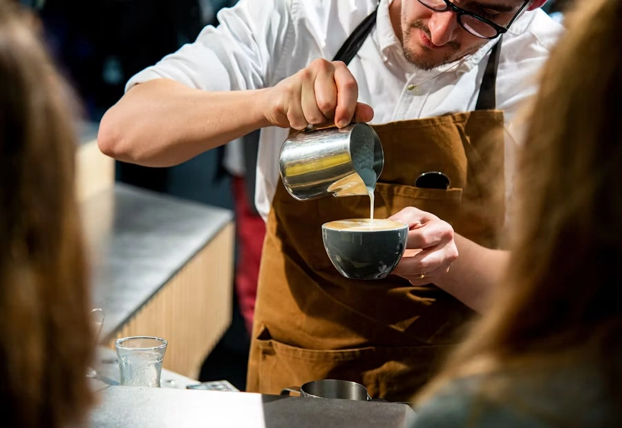 A barista in a brown apron pouring steamed milk into a grey cup at the counter