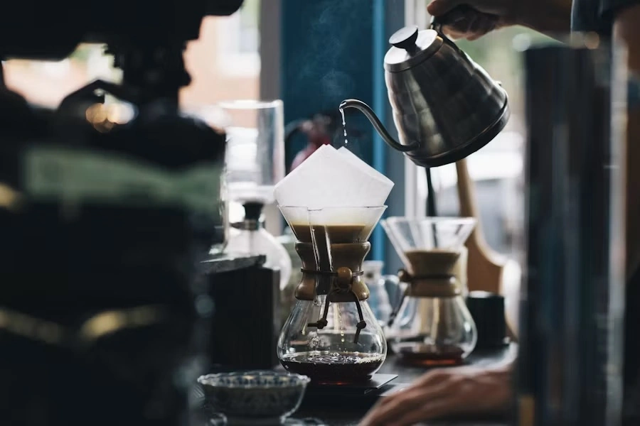 A Chemex pour-over brewing as a kettle drips water over the filter, by a café window