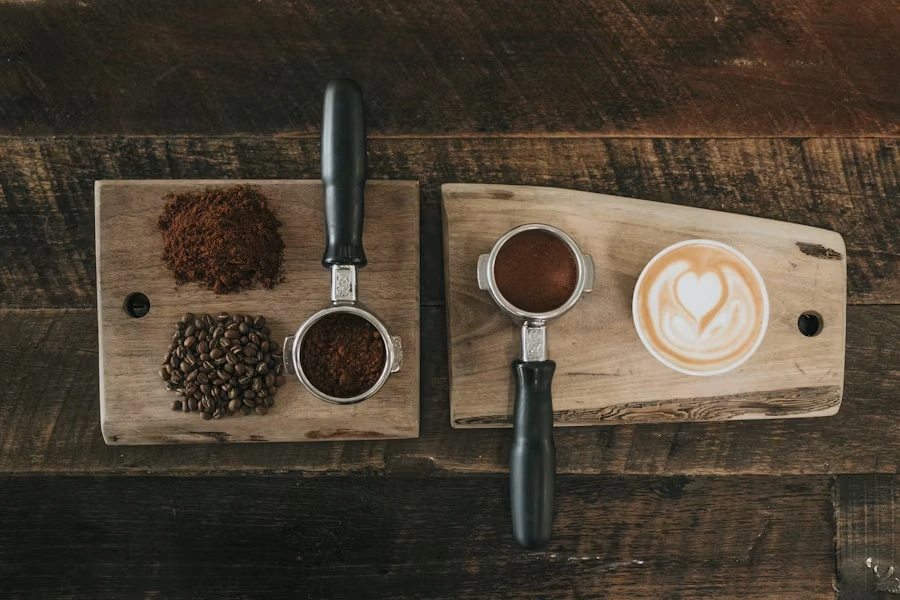 Top-down view of two wooden boards with ground coffee, beans, portafilters and a latte with heart-shaped art