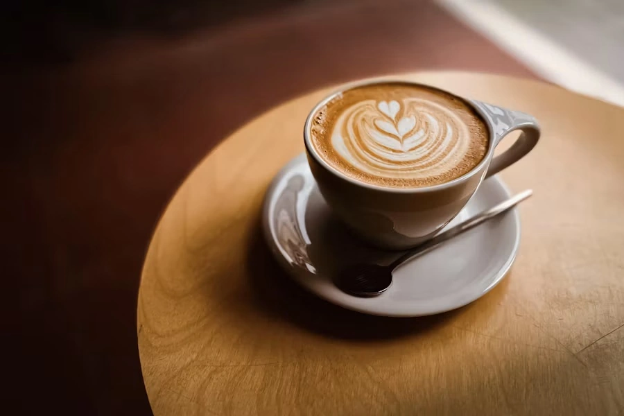 A cappuccino with leaf-shaped latte art in a grey cup on a saucer, on a round wooden table
