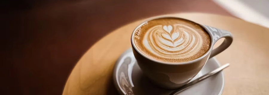 A cappuccino with leaf latte art on a saucer and a wooden table, in a wide crop