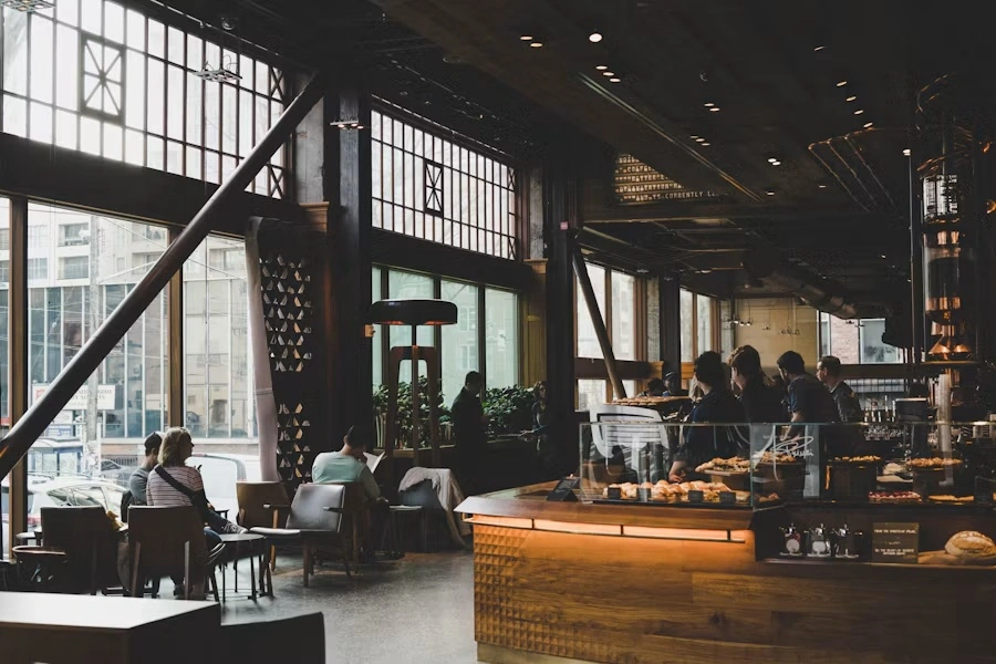 A busy café with tall windows, a warm-lit wooden counter, pastries in a glass case and customers at tables
