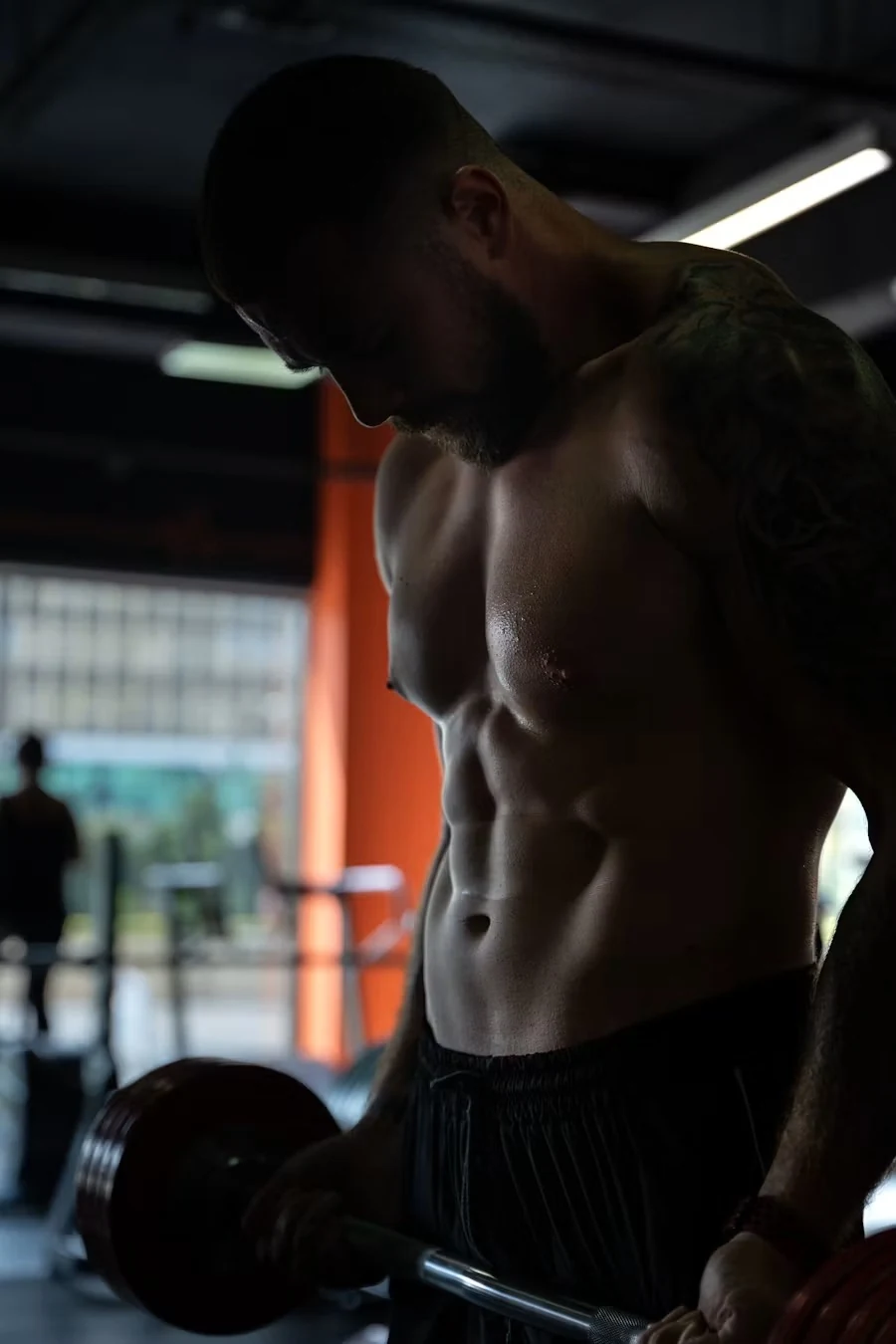 Shirtless lifter looking down while curling a barbell in a dim gym