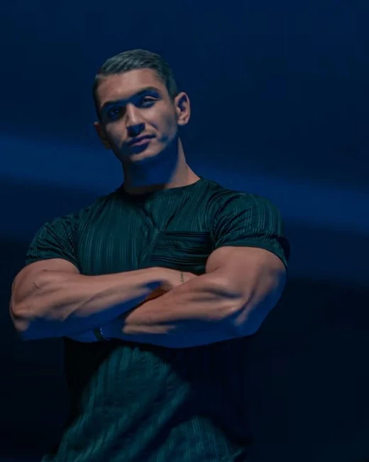 Coach in a dark green t-shirt with arms crossed under blue light