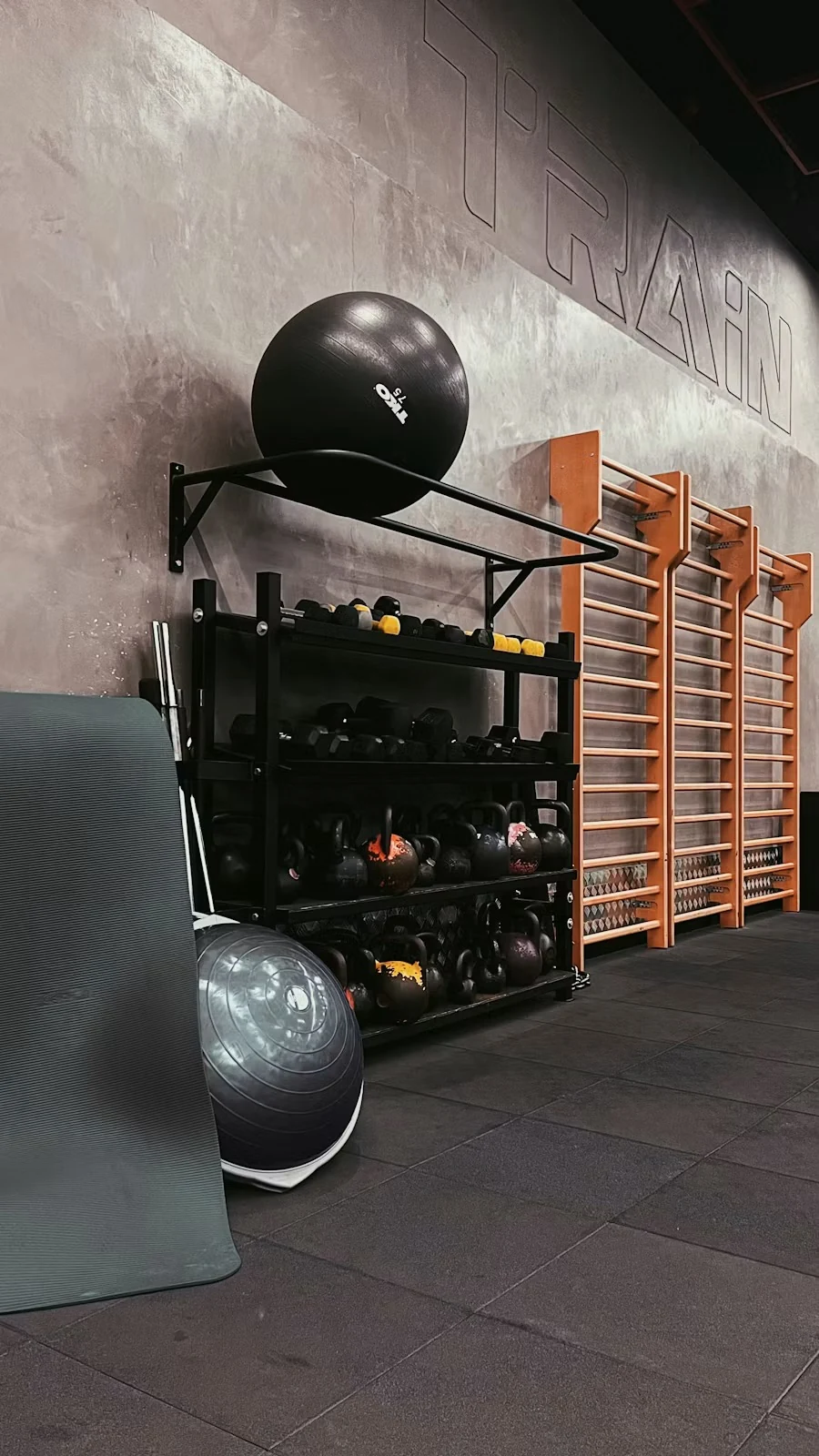 Wall-mounted rack holding an exercise ball, kettlebells and weights beside wooden wall bars