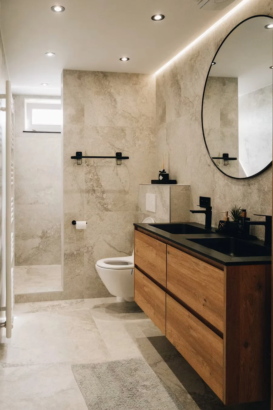 A modern bathroom with a floating timber vanity, black fittings, a round mirror and stone-look tiles