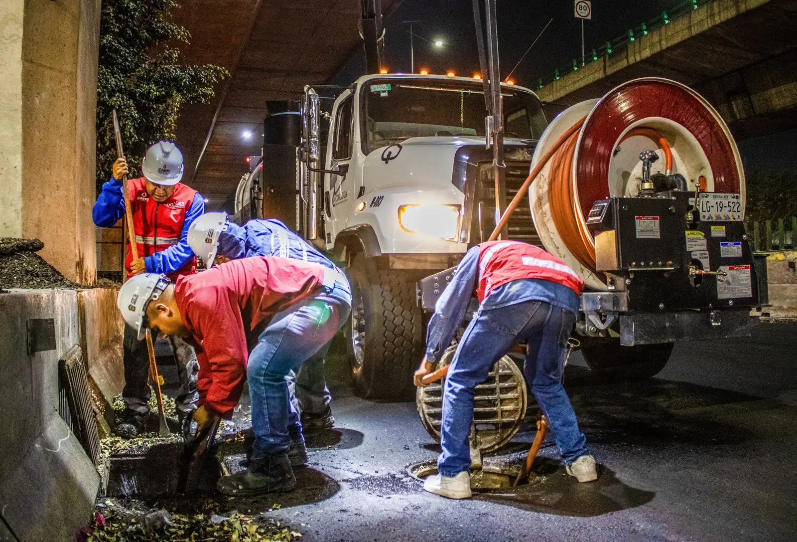 A night crew in hi-vis lifting a manhole cover beside a drain jetting truck with a hose reel