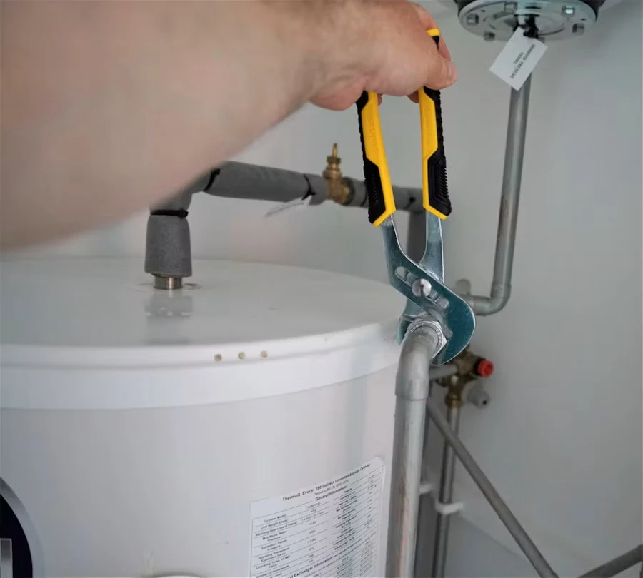 A hand using slip-joint pliers to tighten a fitting on the pipes above a white hot water tank