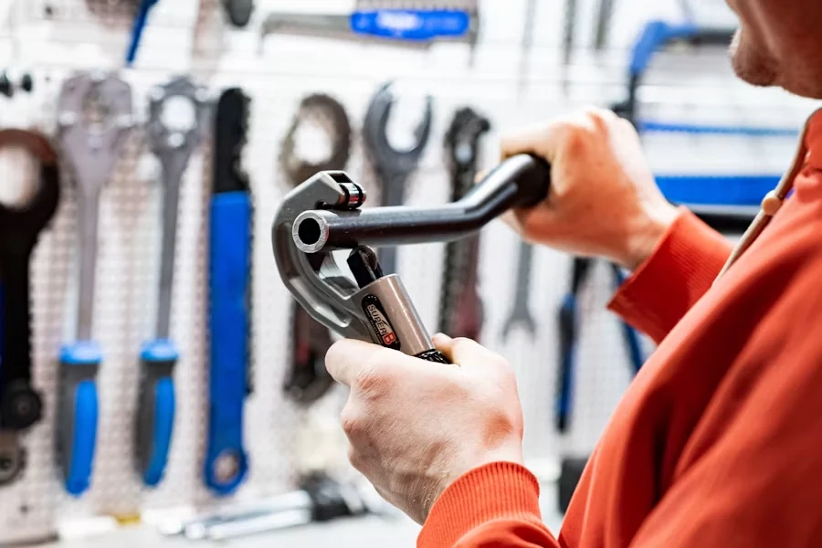 A plumber in an orange jumper holding a pipe cutter in front of a pegboard of wrenches