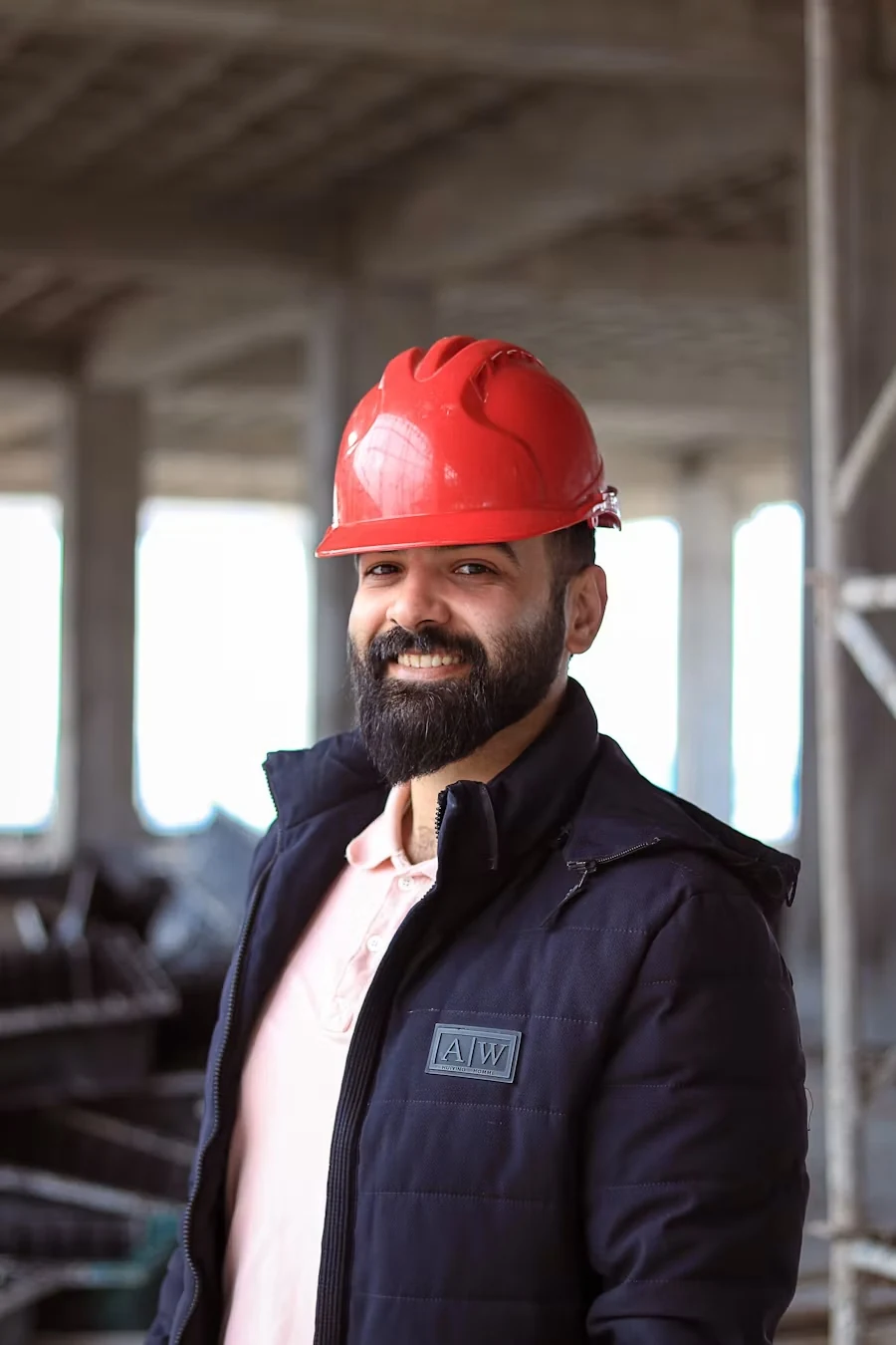 A smiling bearded plumber in a red hard hat and navy jacket standing in a workshop