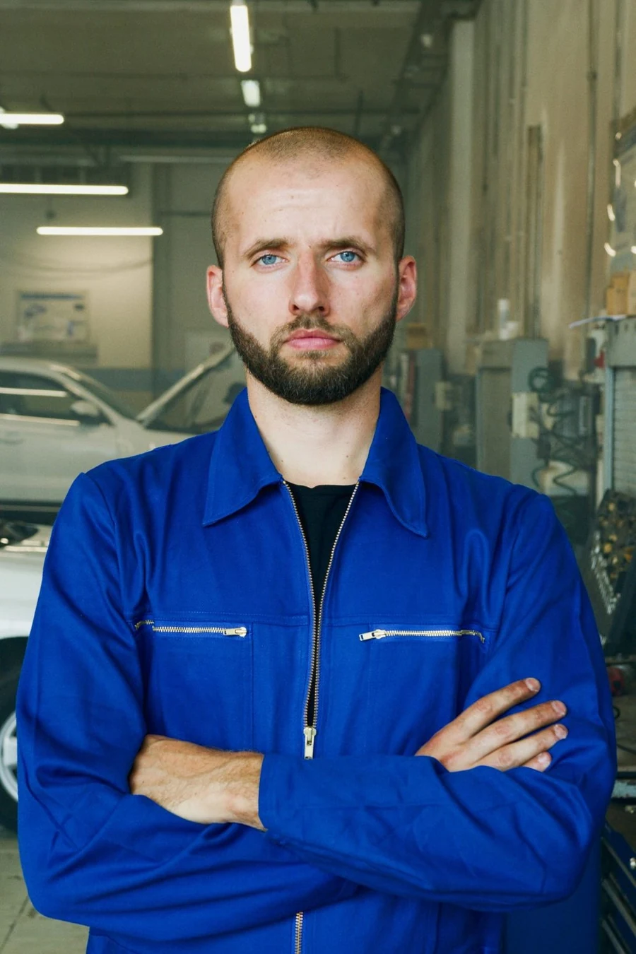 A plumber in a blue work jumpsuit standing with arms crossed in a workshop