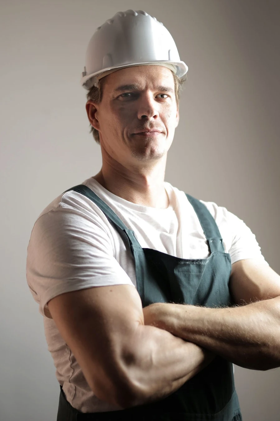 A plumber in a white hard hat and dungarees standing with arms crossed
