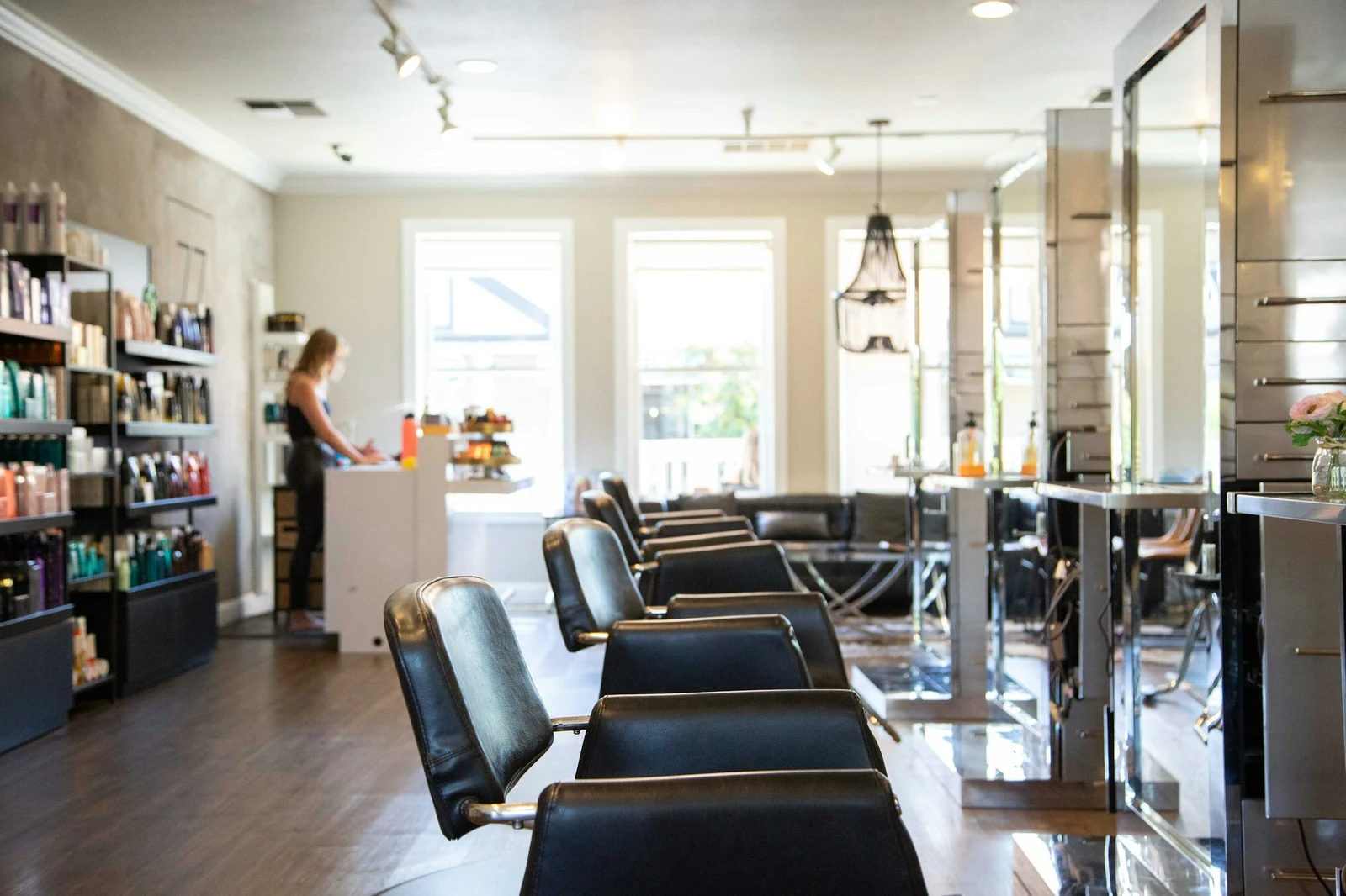 A bright salon with a row of black styling chairs, shelves of products and tall windows